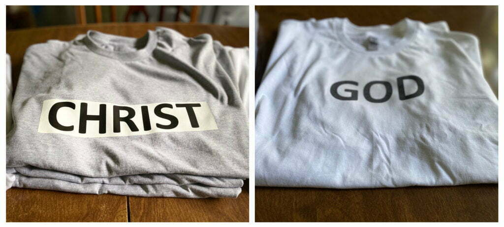 On the left, a gray shirt lays folded on top of a table.  On the top shirt, in bold black text, it reads "CHRIST". On the right, a white shirt lays folded on a table.  On the front of the shirt, in bold black text, it reads "GOD"
