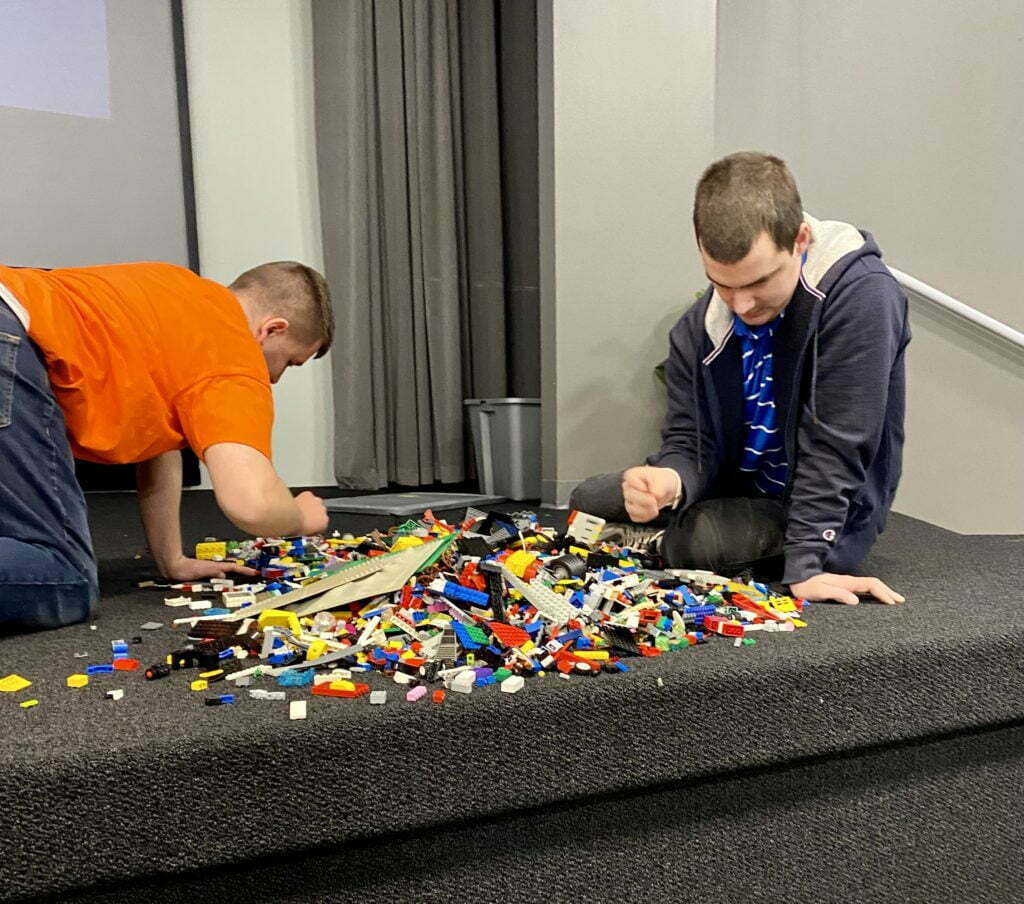 Two men seated on the floor of the classroom stage looking at a large collection of Lego bricks.  The bricks are spread out in a big pile on the floor in front of them.