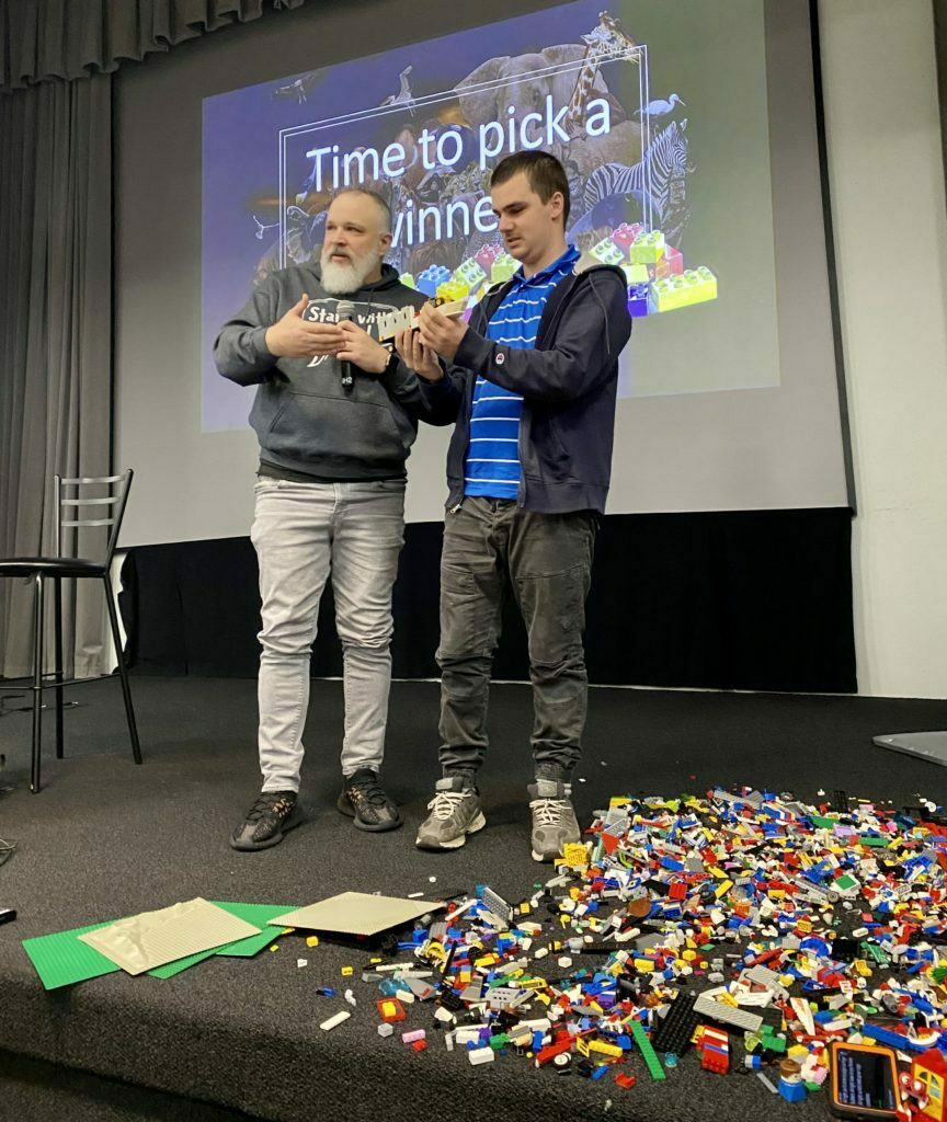 Ryan Wolfe stands to the right of a man  holding a Lego brick creation.  They are both standing on stage in a classroom setting.  Behind them is a large project screen and in front of them on the floor is a large pile of Lego bricks.