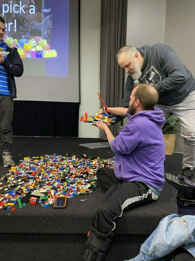 Ryan Wolfe stands on the stage holding a microphone to a man so he can talk to the class.  The man is seated on the floor of the classroom stage and is holding a Lego creation in his hands.  In front of both men, on the floor, is a large pile of Lego bricks.