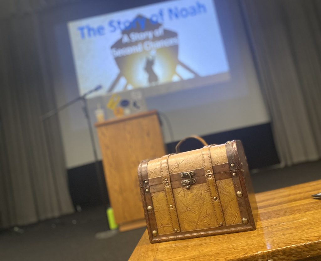 A small brown treasure chest type container sits on top of a table in a classroom setting.  In the background, a large projector screen is positioned behind a podium.