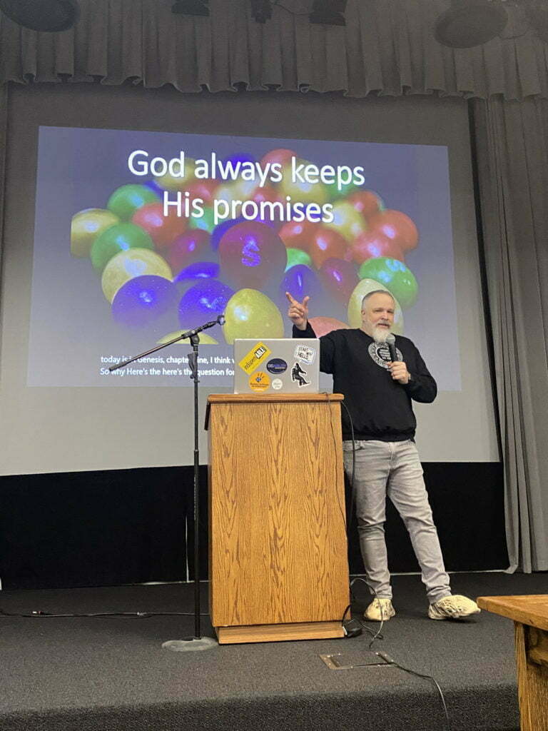 Ryan Wolfe stands in the front of a classroom next to a podium speaking to a group.  Ryan is holding a microphone and raising his hand.  Behind him is a large projection screen that reads: "God always keeps his promises".