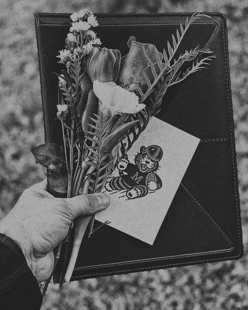 Black and white image of a hand holding a black notebook, small bouquet of flowers and notecard.