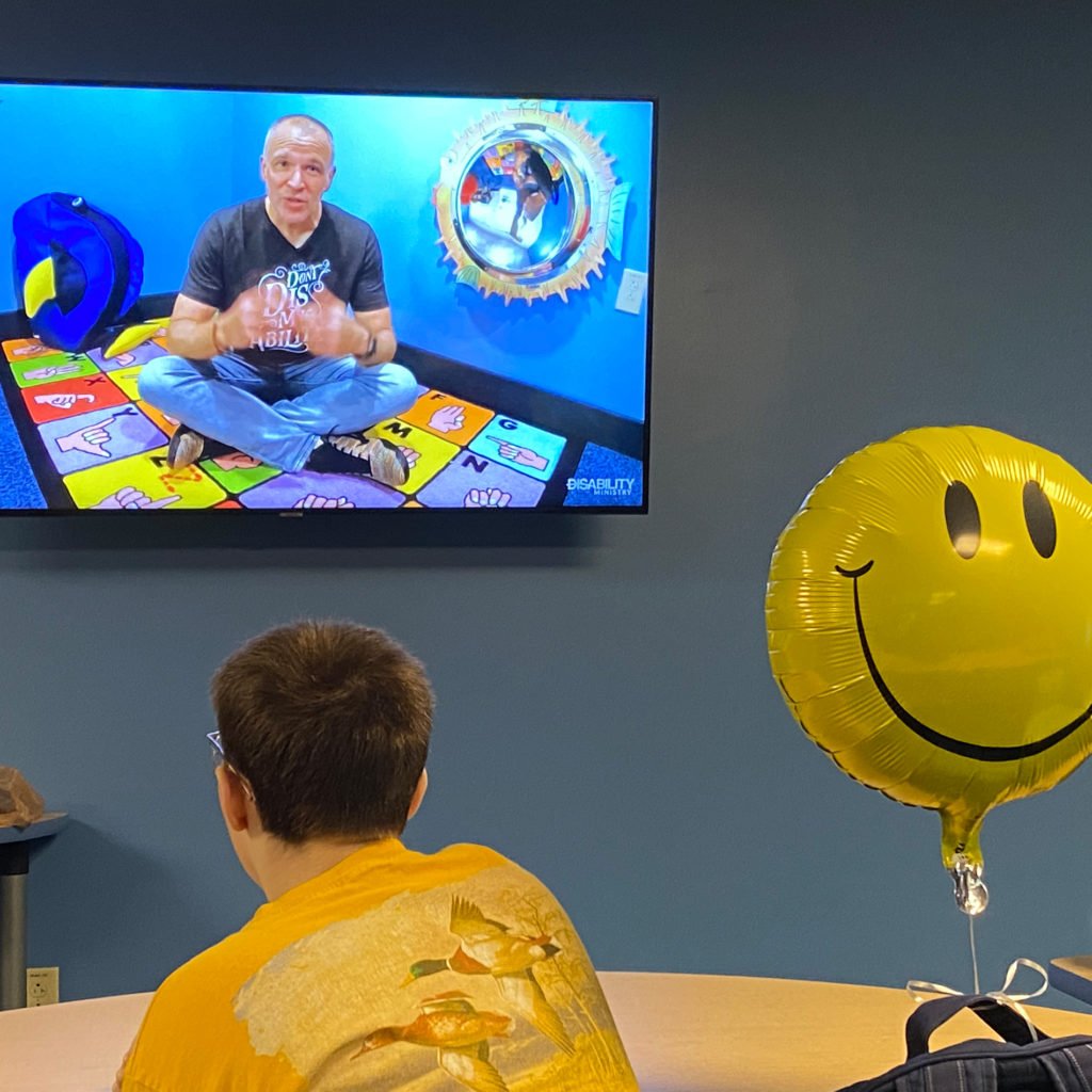 A young man sitting at a desk in the front of a classroom.  He is watching a Sunday School lesson on a wall mounted TV.  To the right of the young man is a yellow smiley face balloon that has been inflated and attached to his backpack.