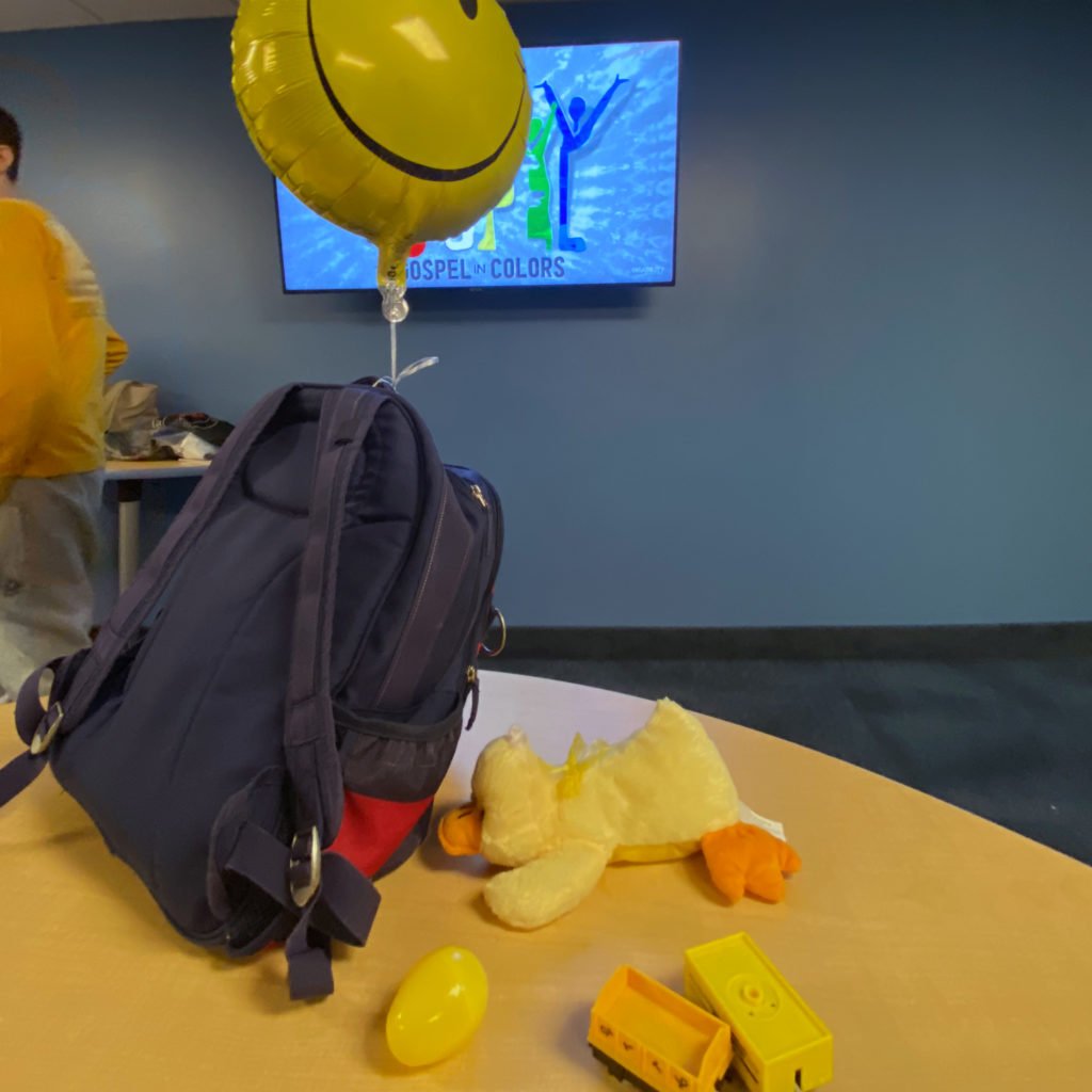 A backpack sits on a table in a classroom.  Attached to the top of the backpack is an inflated yellow smiley face balloon.  On the desk is a plastic yellow egg, stuffed yellow duck, and yellow building blocks.