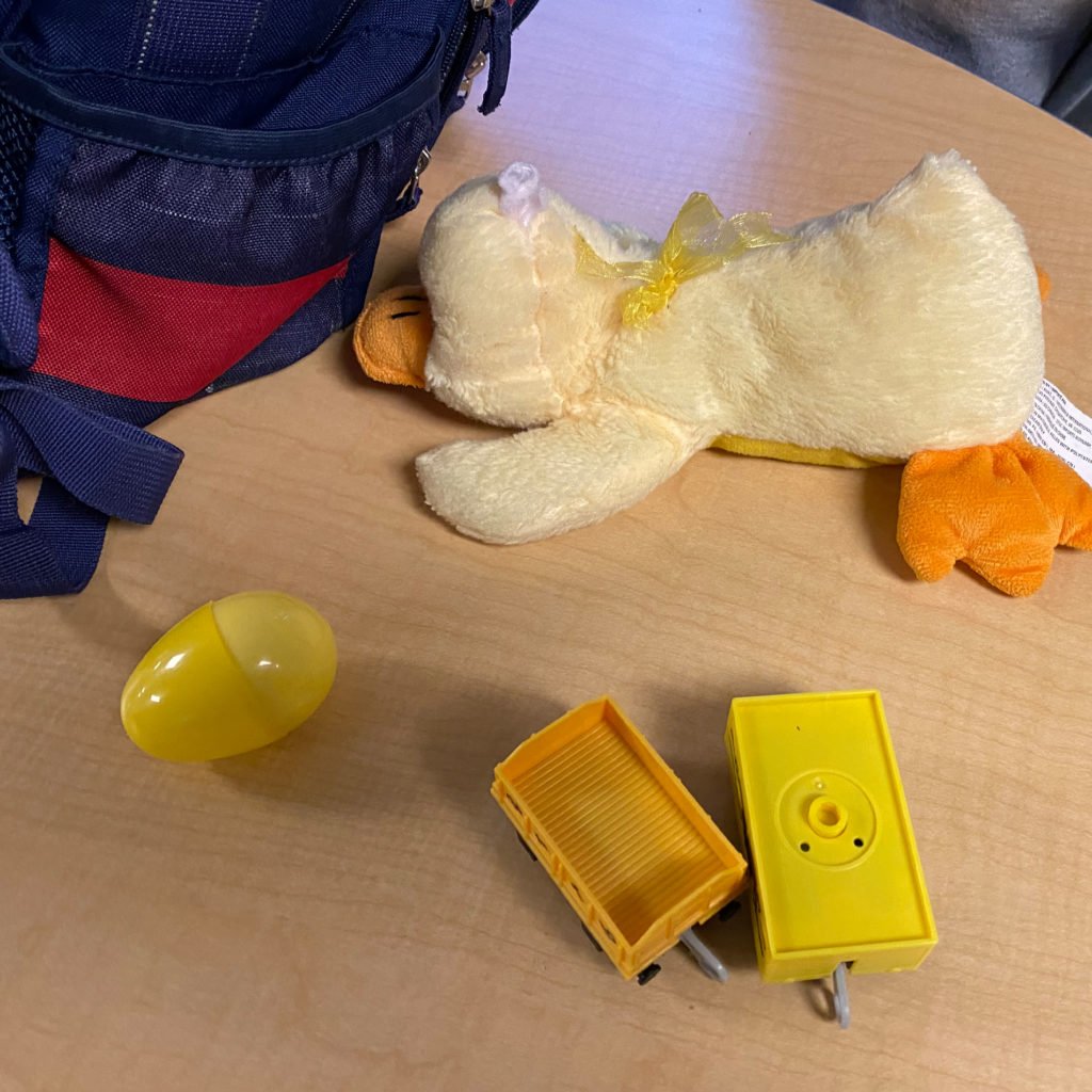 A backpack sits on a table in a classroom.  On the desk is a plastic yellow egg, stuffed yellow duck, and yellow building blocks.