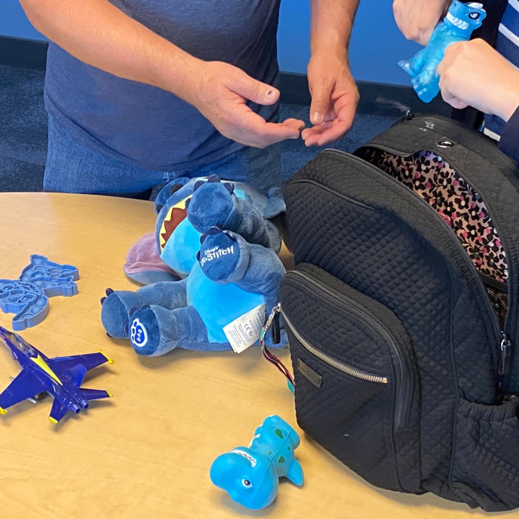 A man and young woman stand next to a desk in a classroom.  The young woman is removing items from the backpack and handing them to the man.  There is a bubble popper sensory item, blue stuffed animal, blue toy airplane, and blue rubber dinosaur.