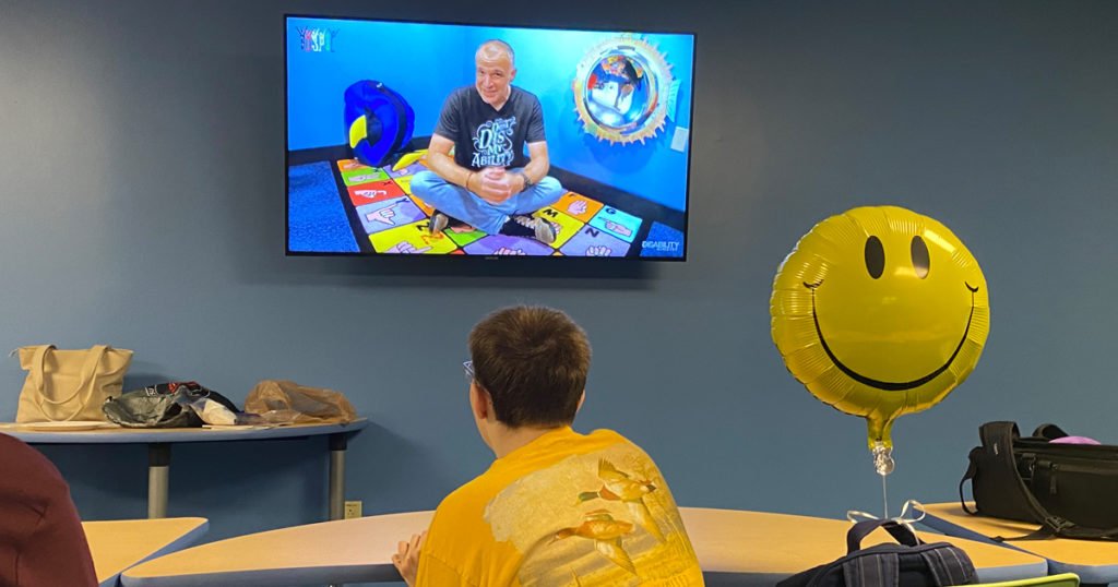 A young man sitting in a classroom watching a lesson on a wall-mounted TV. To the right, next to the young man, is an inflated smiley face balloon.