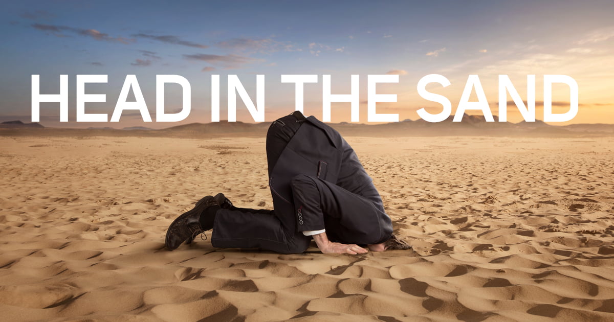 A man wearing a business suit on his hands and knees, with his head stuck entirely in the sand.