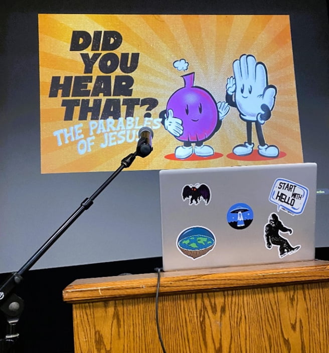 A laptop and microphone sitting on a podium.  In the background, a large screen display is showing an introduction slide for the "Did You Hear That? curriculum series.