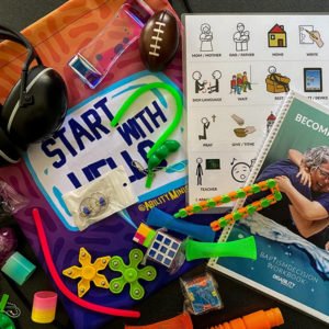 The Disability Ministry Starter Kit on the table features sensory toys and educational materials, including headphones, a football, a book titled BECOME, worksheets, fidget toys, and a colorful cloth bearing the phrase START WITH HELLO, making it perfect for any Disability Ministry.