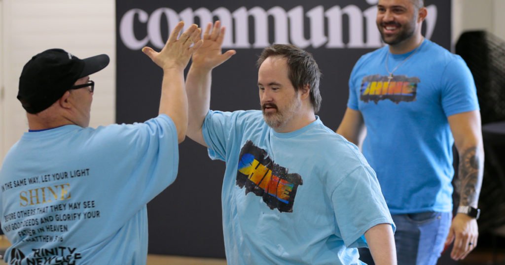 Two men in matching blue t-shirts high-five each other, engaging in nonverbal communication, while a third man in the same shirt smiles in the background. The word community is partially visible behind them.