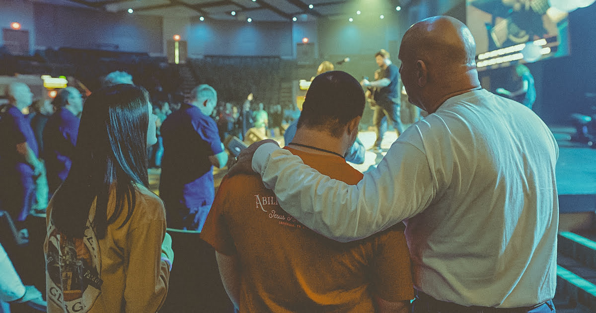 A man places his hand on another person's shoulder amidst a group of people, who are gathered and facing a stage inside a large church auditorium, embodying the essence of worship.