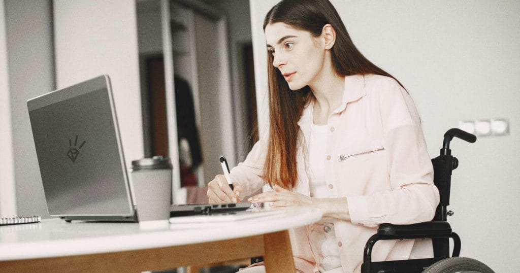 A woman with disabilities sits in a wheelchair, deeply focused on her work at a table. She has her laptop and notepad in front of her and writes thoughtfully with a pen. Her drink is placed beside the laptop as she continues her tasks diligently.