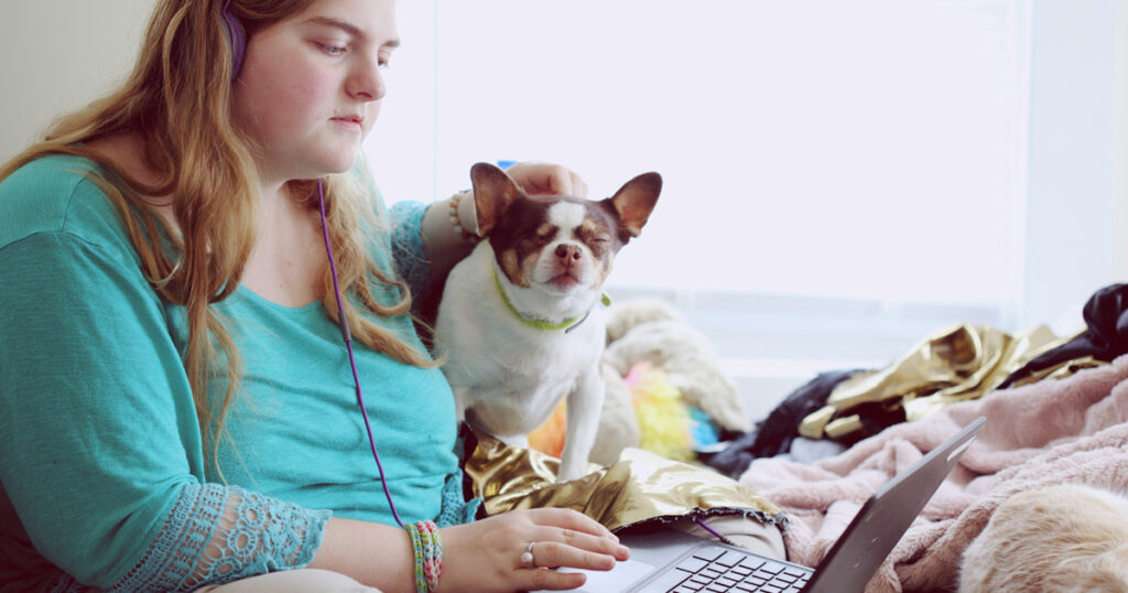 A woman sits on her bed wearing headphones and using a laptop. There is a dog in her lap that she is petting.