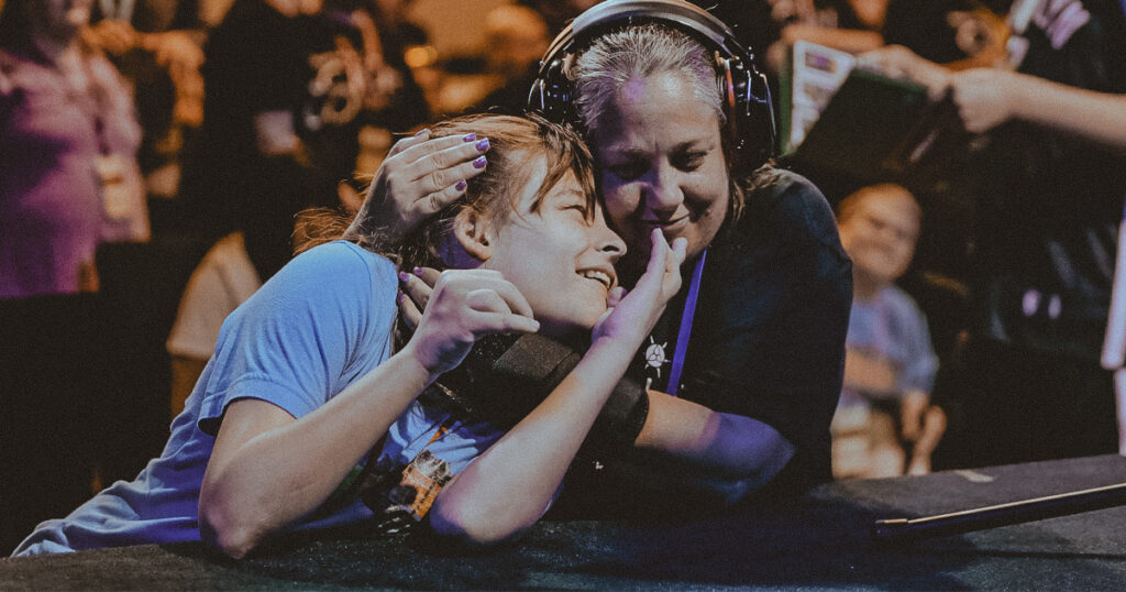 Two people embrace affectionately at an event, one wearing headphones as an ADHD-friendly choice. They are smiling and holding each other close. In the background, a crowd can be seen, highlighting the lively atmosphere of the gathering.