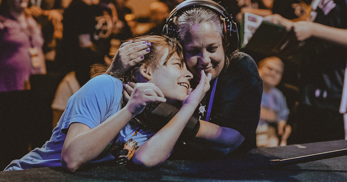 Two people embrace affectionately at an event, one wearing headphones as an ADHD-friendly choice. They are smiling and holding each other close. In the background, a crowd can be seen, highlighting the lively atmosphere of the gathering.