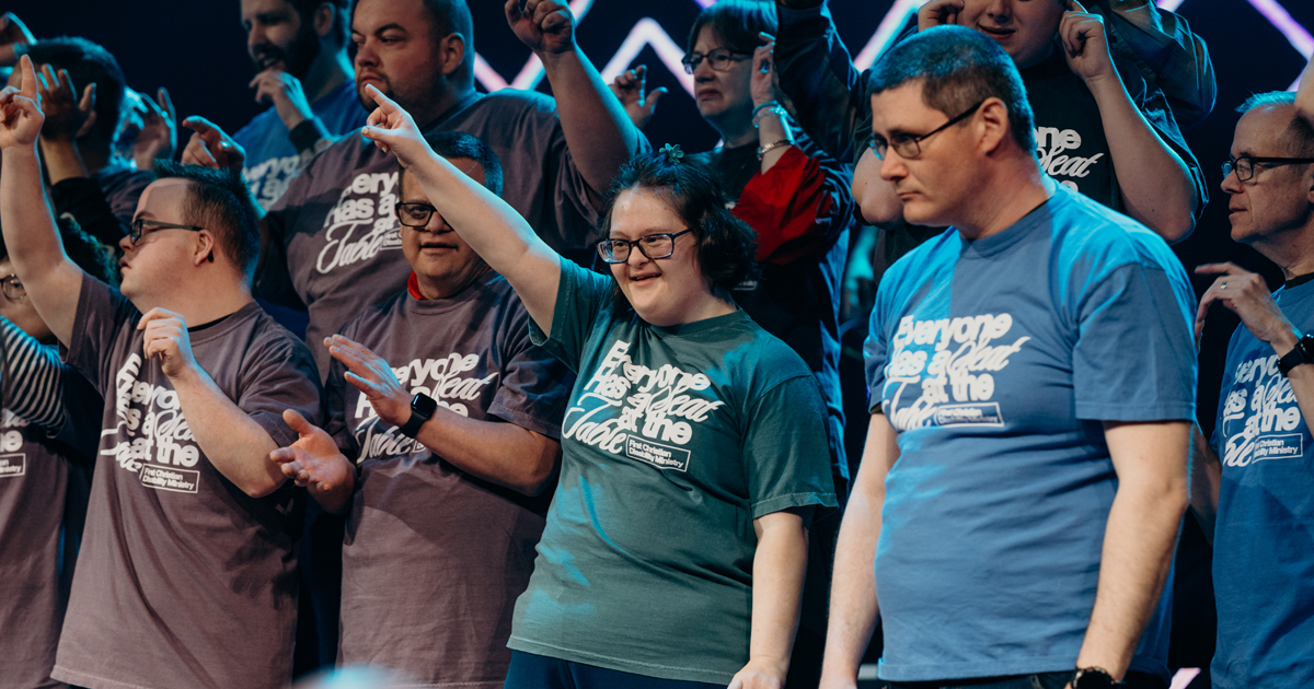 A diverse group of people stands on a well-lit stage, all wearing matching graphic t-shirts. Some members have one arm raised in celebration, expressing joy and unity. The different ages, backgrounds, and personalities of the group highlight the church's commitment to diversity and inclusion as they come together under vibrant stage lighting.