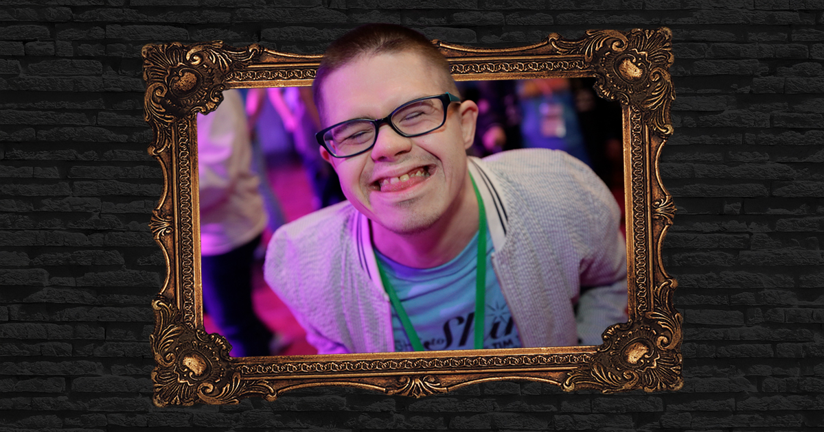 A person with glasses smiles warmly at the camera, their friendly expression illuminated softly. They wear a light sweater and a lanyard draped neatly around their neck, suggesting a casual yet purposeful presence. The portrait is elegantly framed by an ornate gold frame rich with intricate detailing, lending the image an air of classical importance. Behind this gilded boundary, the backdrop consists of dark, textural brickwork in subdued shadow—enhancing both the subject’s prominence and the sense that this is an unexpected hidden masterpiece on display.
