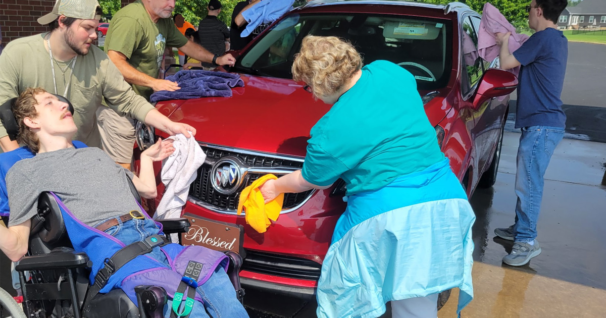A group of people, including one in a wheelchair, washing a red Buick together outside on a sunny day, showing church inclusivity.