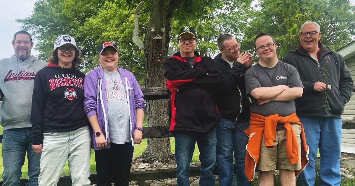 Seven adults stand outdoors by a tree and fence on a cloudy day, smiling—ideal to showcase your Ministry with Adults or welcoming group.