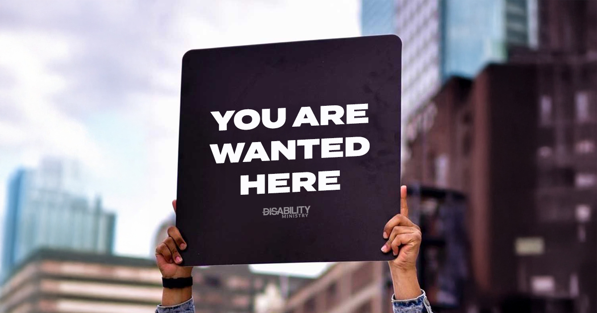 A person's hands are shown holding a large sign that reads You are wanted here with a city backdrop
