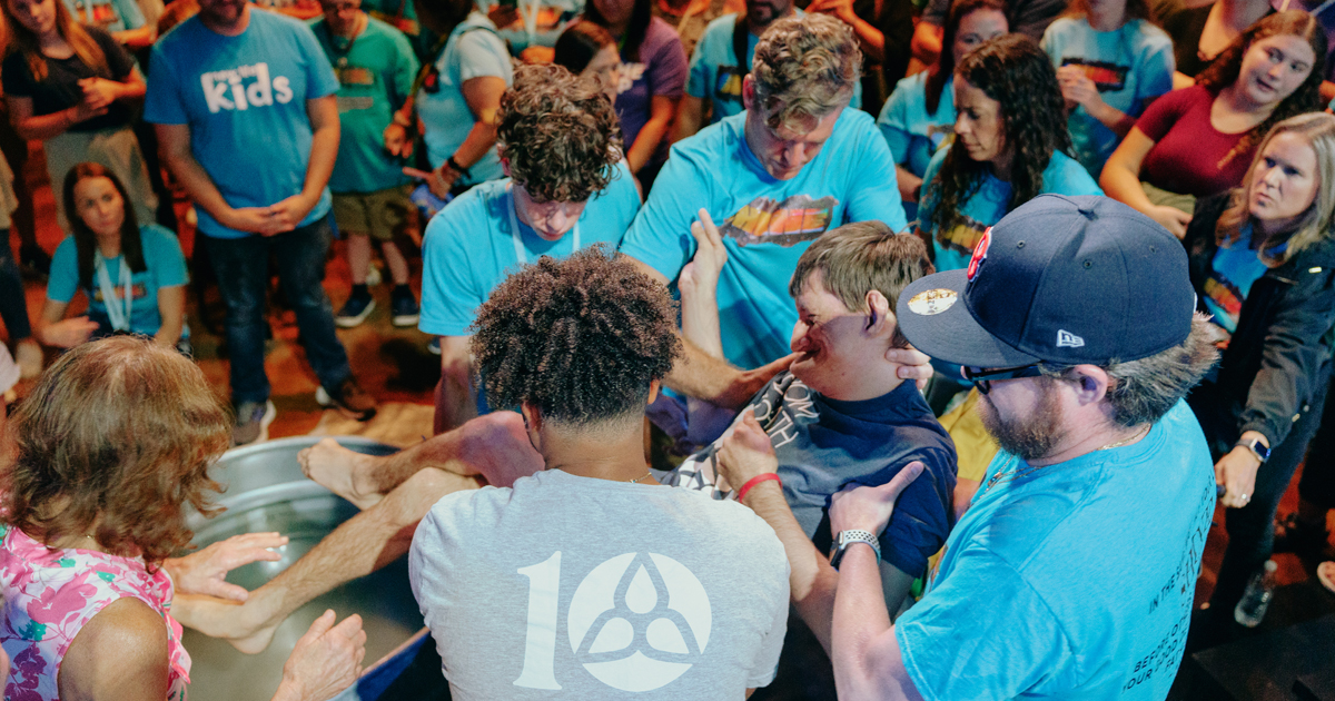 People help a boy being baptized in a water tub, surrounded by others in blue shirts, evoking ministry and John’s Baptism.