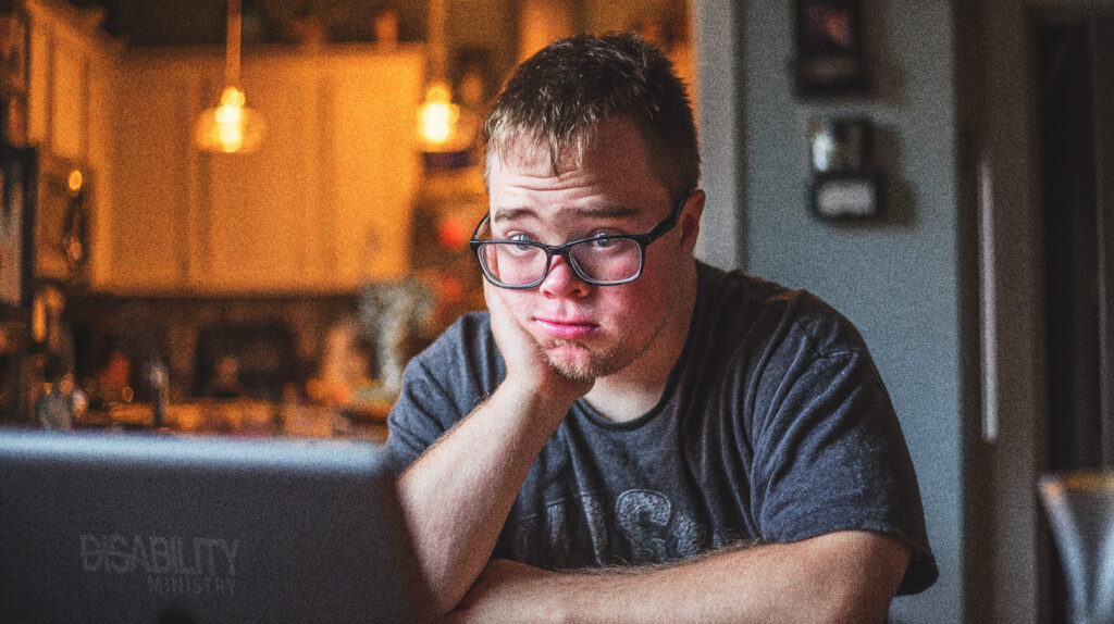 A young adult male is sitting at his kitchen table looking at his laptop.  He has a sad expression on his face.