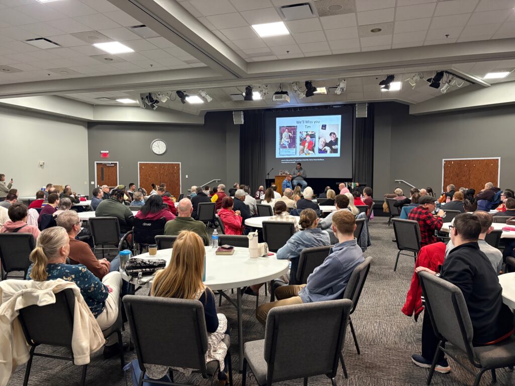 Attendees sit at round tables in a conference room, watching a presentation on grief and loss shown on a screen up front.