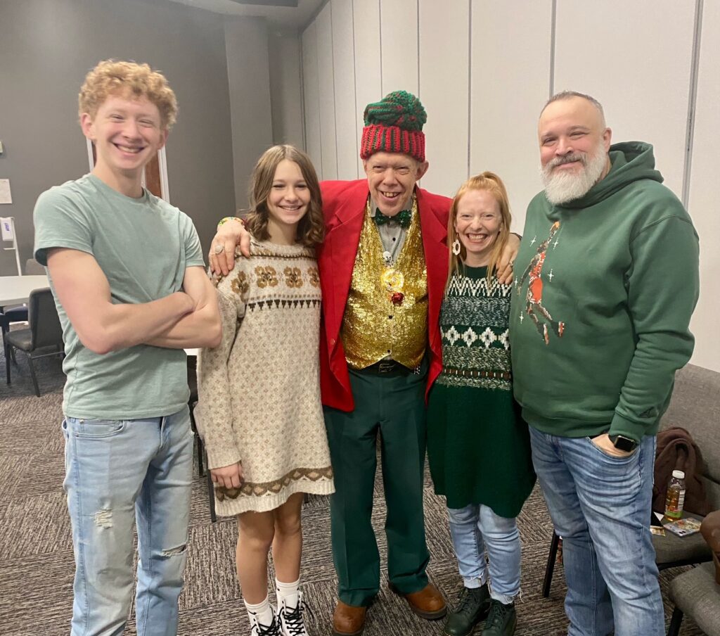 Five people stand indoors, smiling at the camera. The person in the center is in festive attire; others wear warm casual clothes, expressing unity even through loss.