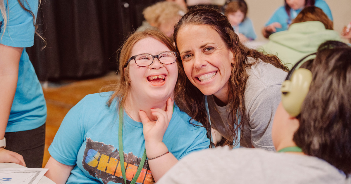 Two people beam at the camera&mdash;a young girl in glasses and a blue shirt, and an adult woman with brown hair&mdash;radiating belonging together. Other people are visible behind them.