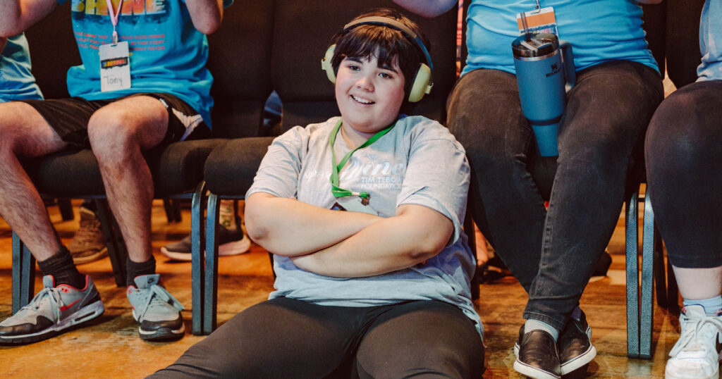 A person with headphones sits cross-legged on the floor before a group, smiling and relaxed at an inclusive indoor event.