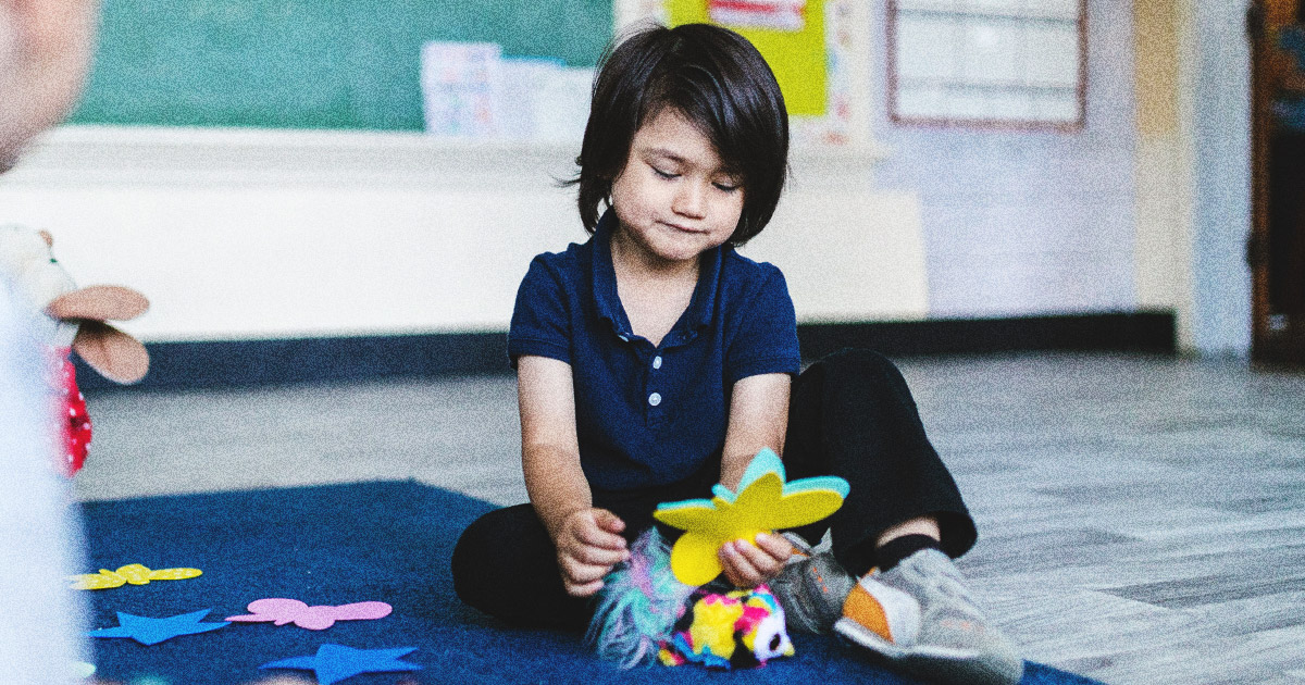 A child sits cross-legged on the floor in a classroom, holding a yellow paper cutout amid colorful shapes, encouraging creativity.
