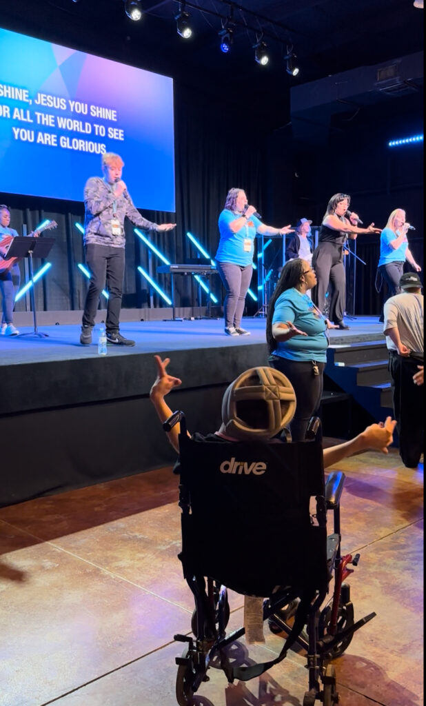 A child in a wheelchair observes a worship band performing on a church stage, as multiple singers’ voices blend harmoniously and song lyrics are displayed on a screen behind them.