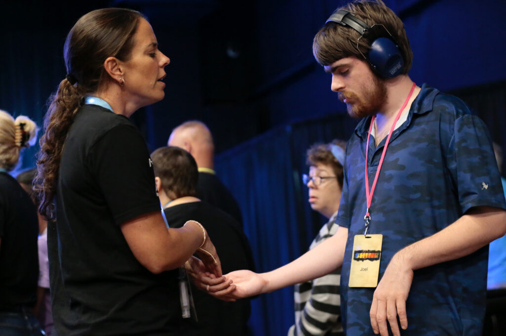 A woman clasping the hand of a young man with headphones and a name tag expresses unity in diversity. Several other people in the background capture the inclusive atmosphere of Different Voices, One Worship.