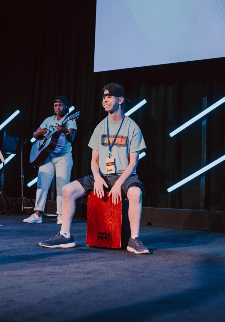 Two performers are on stage; one strums a guitar, and the other sits on and plays a red cajón percussion instrument. Both are dressed casually with hats, illuminated by vibrant stage lighting behind them. Their collaboration embodies Different Voices united in One Worship experience.