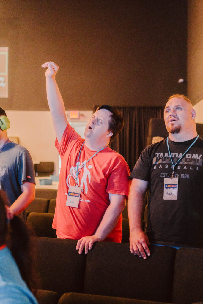 A man wearing a red shirt and a name tag lifts his arm as he sings during worship, standing beside another man in a black shirt among rows of chairs, their voices blending harmoniously.