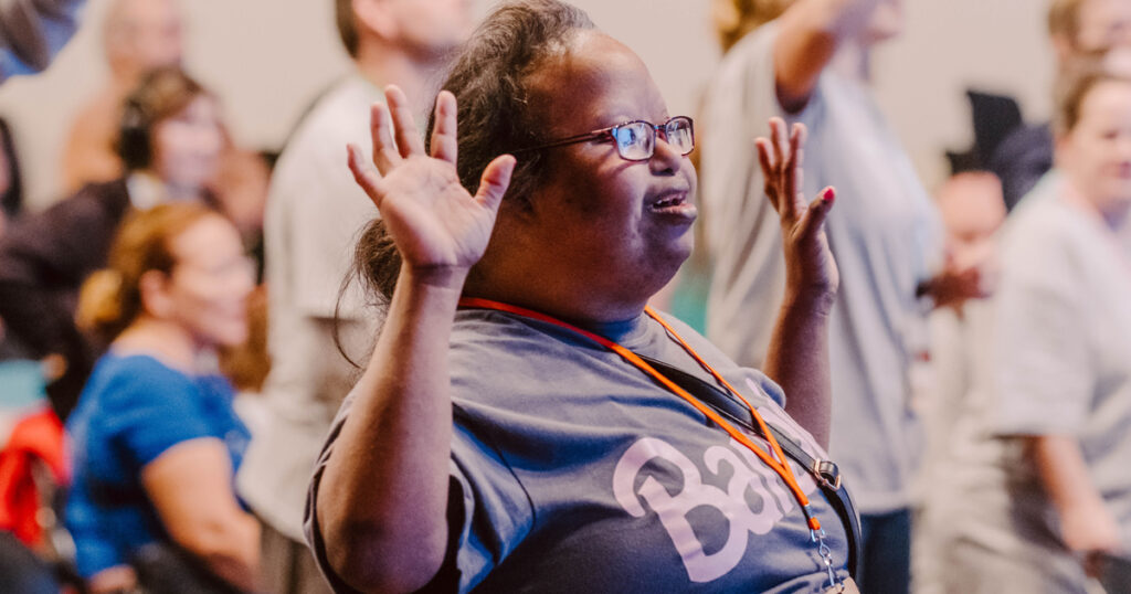 A woman wearing glasses stands among a group of people at an indoor event, lifting her hands in worship.