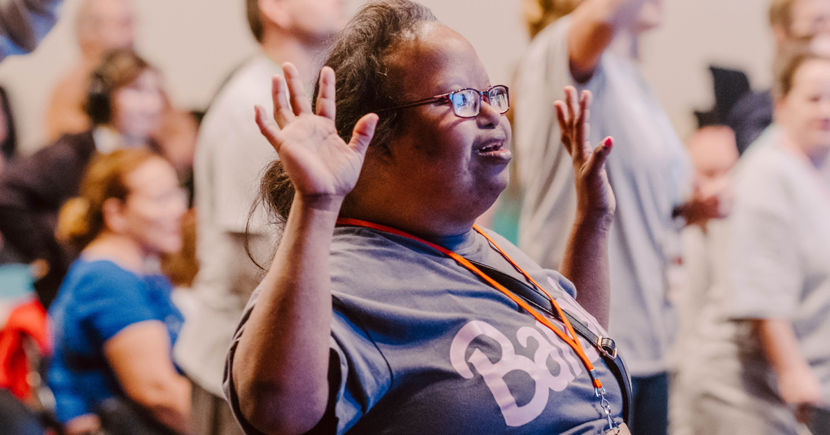 A woman wearing glasses stands among a group of people at an indoor event, lifting her hands in worship.