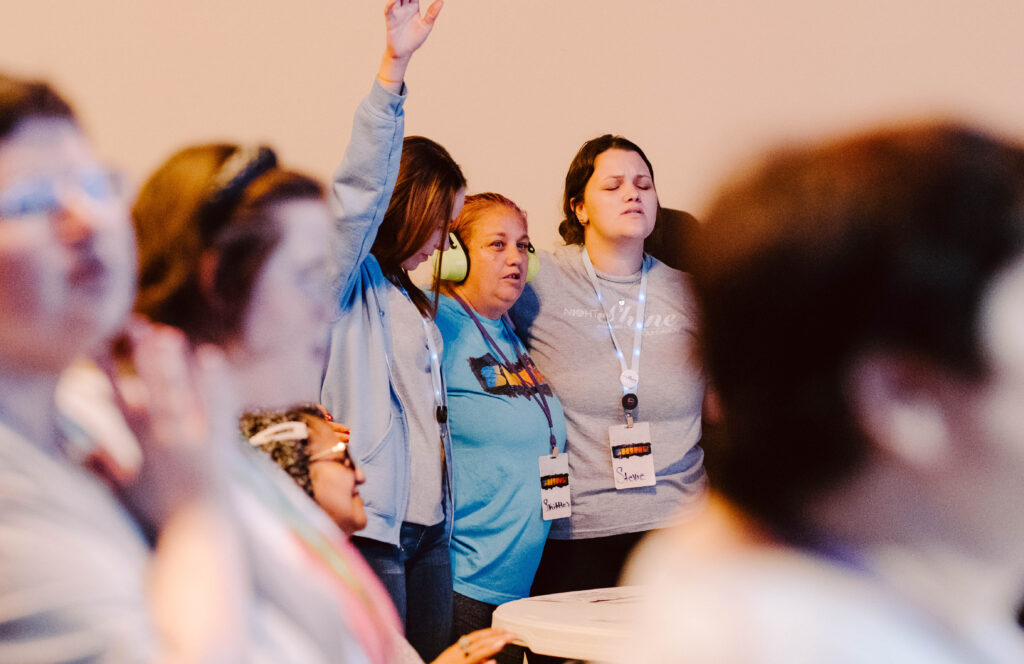 A group of people gather closely inside, some with eyes closed and one raising a hand, together in One Worship. Many wear name tags and headphones, symbolizing the unity of Different Voices in a collective experience.