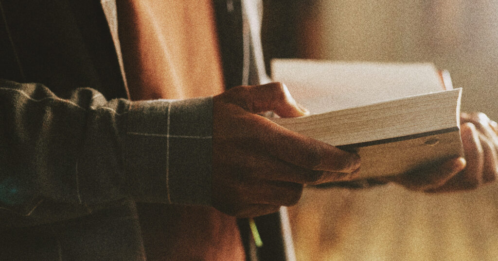 An elderly individual, dressed in a long-sleeved shirt or jacket, sits under gentle lighting while flipping through the pages of an open book.