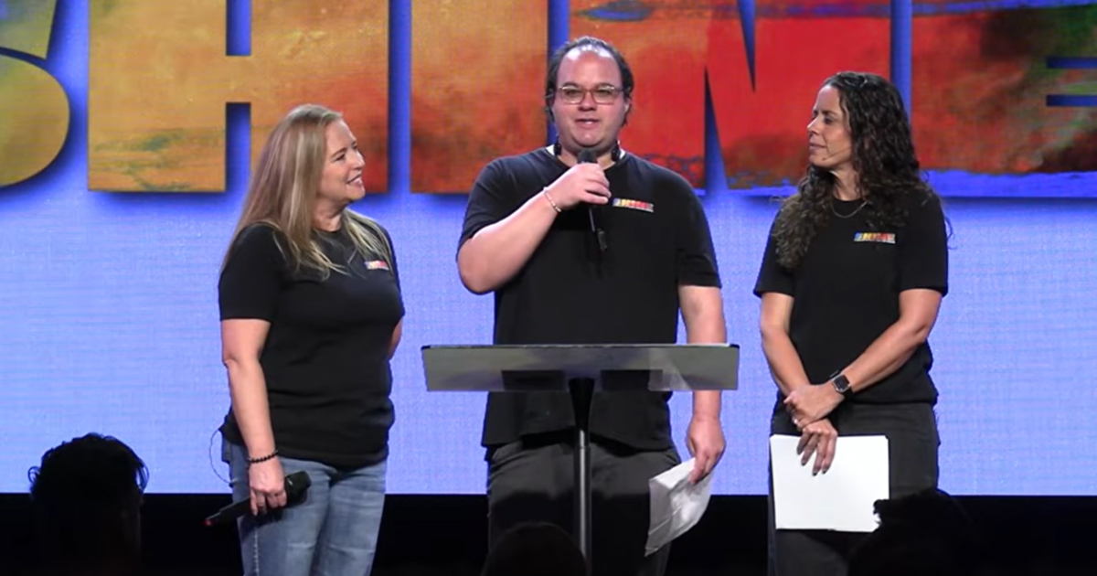 Three people stand on stage behind a podium, all dressed in black shirts, with a vibrant SHINE sign hanging behind them. One person is speaking into a microphone, discussing their church&rsquo;s special needs ministry.
