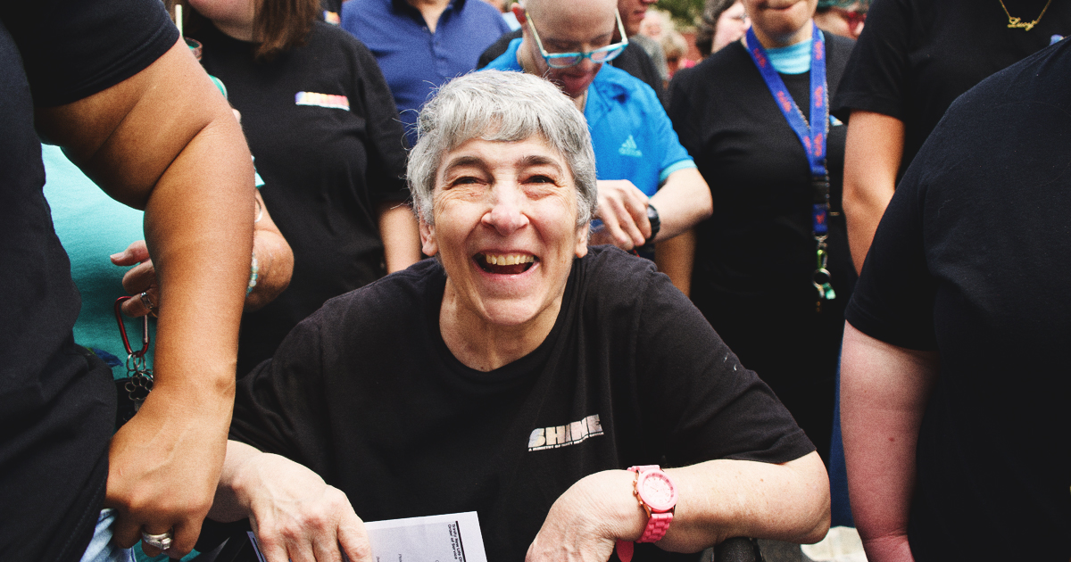 A cheerful older woman with short gray hair sits amidst a crowd, clutching papers to her chest. She is dressed in a black shirt and sports a pink watch that perfectly complements her look. People stand nearby, several of them wearing lanyards and blue shirts.